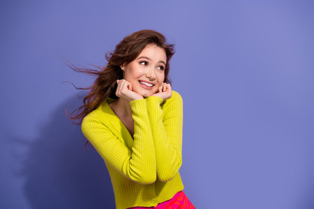 Portrait of a cheerful young woman in a yellow cardigan on a vibrant purple background, expressing...