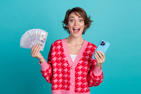 Photo of pretty impressed lady wear red cardigan winning modern gadget holding money fan isolated turquoise color backgroundの写真素材