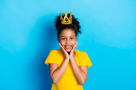 Cheerful young girl with a golden crown in bright yellow attire against a vibrant blue background, expressing excitementの写真素材