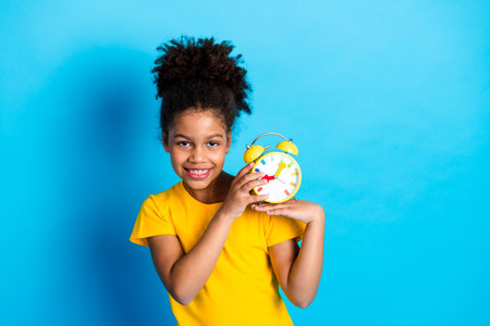 Cheerful young girl holding a vibrant clock, dressed in stylish casual yellow shirt with joyful...