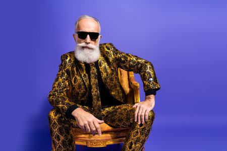 Charismatic senior man in eccentric snake print suit and sunglasses seated confidently against a...