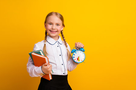 Smiling schoolgirl holding book and an alarm clock against a cheerful yellow background, promoting education and learningの写真素材