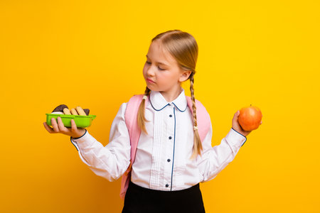 Smiling schoolgirl holding an apple and snacks, pondering a choice amid yellow background, education and healthの写真素材