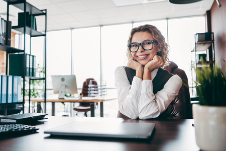 Young businesswoman smiling at her desk in a modern corporate office surrounded by workspace interior design elementsの写真素材
