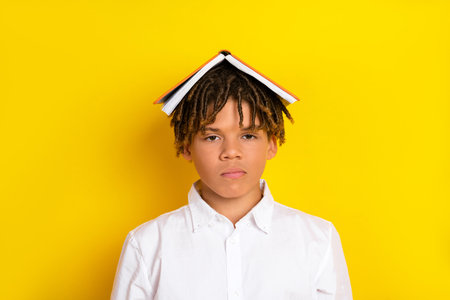 Young student in white shirt with book on head against yellow background, representing education and...
