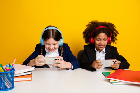 Two cheerful schoolgirls play games together on phones, wearing headphones and sitting at a desk with supplies in a classroom.の写真素材