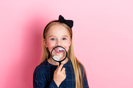 Adorable young girl holding a magnifying glass playfully on a vibrant pink background, radiating...