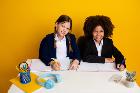Two schoolgirls studying together in a classroom setting with bright yellow background and school supplies showcasing educationの写真素材