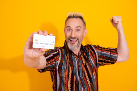 Charismatic man happily showcases a credit card against a yellow background, exuding confidence and...
