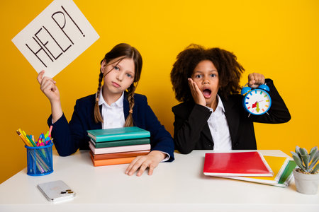 Two schoolgirls in uniforms with books, clock, and help sign at desk on bright yellow background expressing urgencyの写真素材