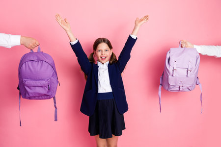 Charming schoolgirl celebrating school achievements with arms raised surrounded by vibrant purple backpacks on a pink backdropの写真素材