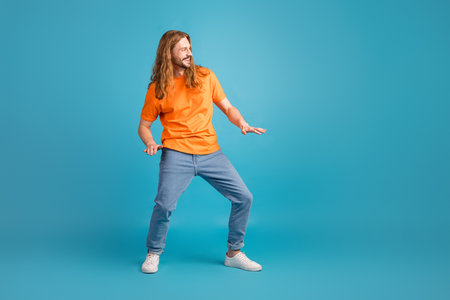 Young man in vibrant orange t-shirt making playful gestures on blue background, showcasing trendy casual fashionの写真素材