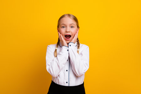 Happy schoolgirl in uniform expressing surprise with hands on cheeks against yellow background, showcasing cheerful childhoodの写真素材