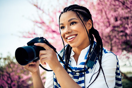 Young woman holding a camera outdoors surrounded by blooming flowers during a sunny spring dayの写真素材