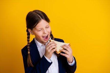 Cheerful preteen schoolgirl with pigtails enjoying a sandwich against a vibrant yellow backgroundの写真素材