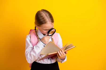 Young girl in school uniform holding a book and magnifying glass while smiling on a bright yellow backgroundの写真素材