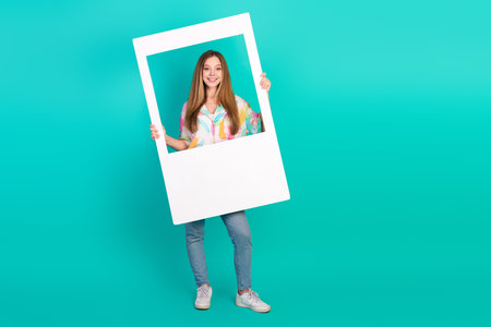 Stylish Young Woman Holding a Picture Frame on a Turquoise Background in Casual Outfitの写真素材