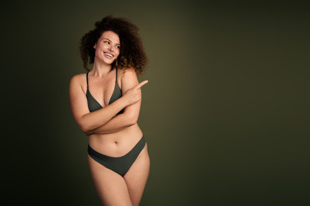 Confident Young Woman Embracing Natural Beauty in Stylish Dark Green Lingerie Against a Solid Colored Studio Backgroundの写真素材