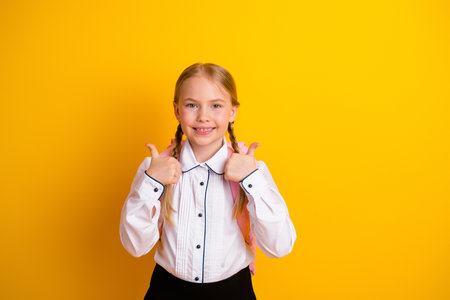 Smiling schoolgirl in formal uniform giving thumbs up against a bright yellow background, showcasing...