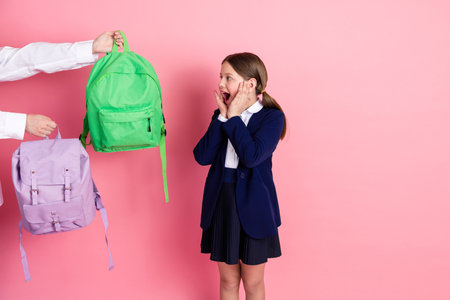 Excited schoolgirl in formal uniform reacting to colorful backpacks against a pink background for...