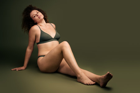 Young confident woman with curly hair in stylish lingerie on a khaki background, embracing self acceptanceの写真素材