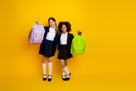 Cheerful schoolgirls in uniform with vibrant backpacks posing on a bright yellow background, expressing friendship and school spiritの写真素材