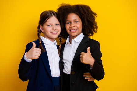 Diverse young friends in professional attire smiling together holding thumbs up on vibrant yellow backgroundの写真素材