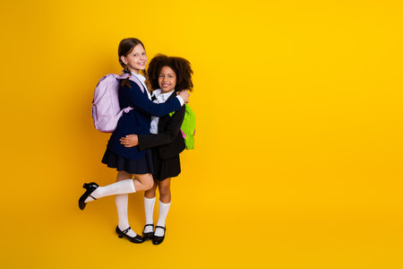 Two young schoolgirls in stylish uniforms posing with colorful backpacks on a vivid yellow background, expressing happinessの写真素材