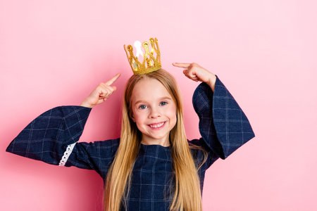 Happy young girl posing with golden crown accessory on pink background enjoying playful creative and cheerful momentの写真素材