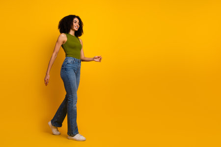 Young woman with curly hair in casual outfit walking confidently against a yellow background, showcasing vibrant style and charmの写真素材