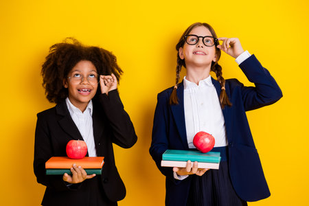 Two schoolgirls in uniforms holding books and apples against a vibrant yellow backdrop, expressing joy and togethernessの写真素材