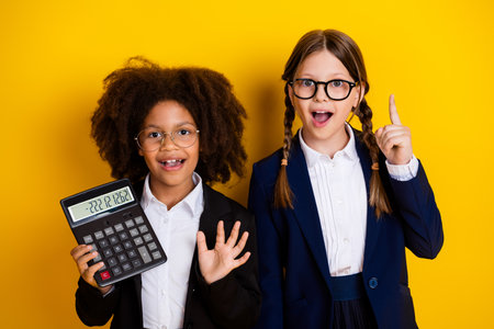 Two excited schoolgirls wearing uniforms holding a calculator and gesturing, standing against a vibrant yellow backgroundの写真素材