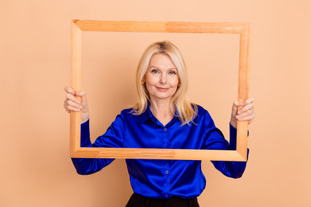 Mature Businesswoman Holding a Wooden Frame in Hands Over a Beige Background in a Stylish Blue Shirt