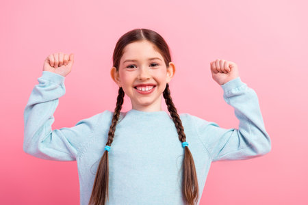 Cheerful young girl with braided hair raising her arms in a pink backdrop showcasing happiness and positivityの写真素材
