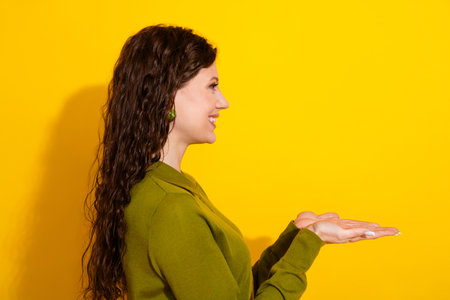 Side profile of a cheerful young woman presenting a gesture on a vibrant yellow background wearing a green sweaterの写真素材