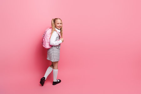Happy young schoolgirl in uniform with backpack, standing on a pink background, representing education and knowledgeの写真素材