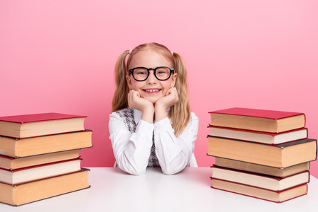 Smiling young schoolgirl sitting at a desk surrounded by book against a pink background symbolizing education and knowledgeの写真素材