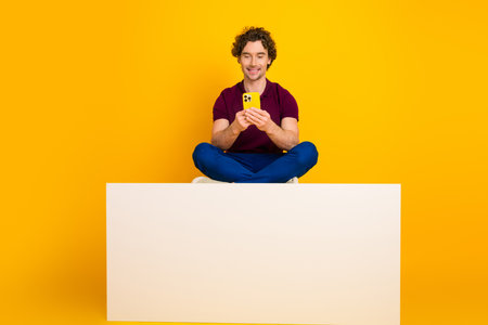 Young man with curly hair sitting cross-legged, holding a yellow phone against a bright yellow...
