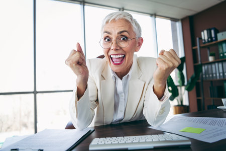 Positive senior businesswoman celebrating success at workplace desk in tidy office environment with enthusiasmの写真素材