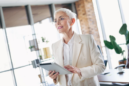 Mature professional woman confidently working on a tablet in a modern office environment with a bright outlookの写真素材