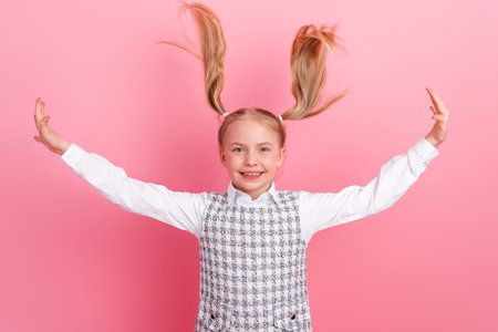 Preteen girl with blonde ponytails happily posing in formal school uniform on solid pink background showing playful gesturesの写真素材