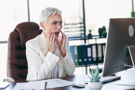 Elegant senior businesswoman expressing surprise while working in a professional office environment with a computerの写真素材
