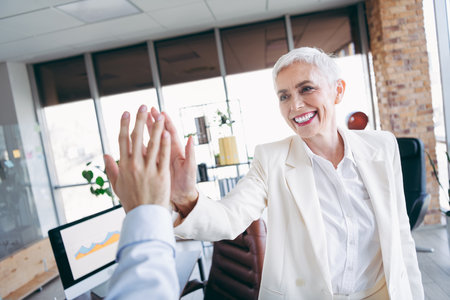 Smiling senior businesswoman in formal white attire giving a professional high five in an office settingの写真素材