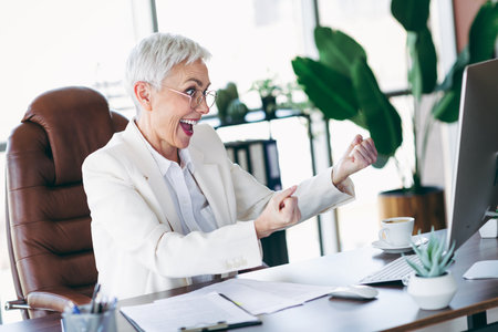 Happy senior businesswoman celebrating success at her office workspace while working on a projectの写真素材
