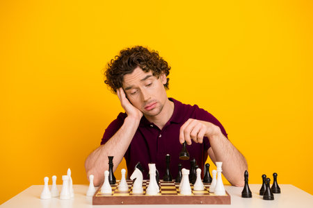 Young man playing chess while showing a reflective expression sitting at a table against a bright yellow backgroundの写真素材