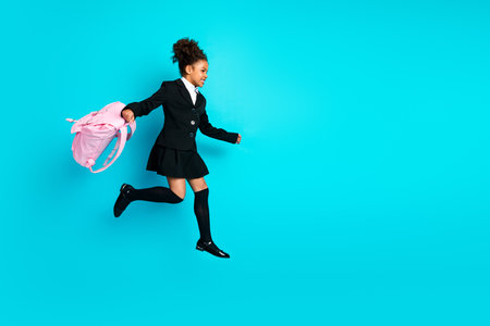 Young schoolgirl in stylish uniform jumping with pink backpack on vibrant turquoise background full of happinessの写真素材