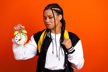 Young student with braids holds yellow alarm clock, looking worried against orange background in...
