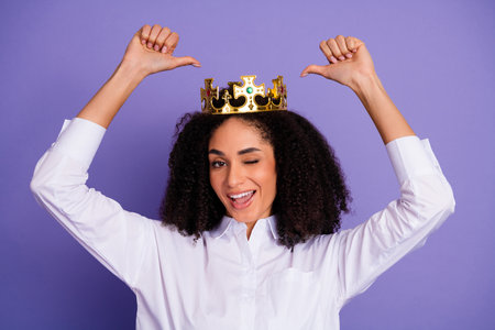 Young woman wearing a crown giving a playful gesture on a vibrant purple backgroundの写真素材