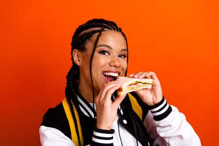 Young woman enjoying a fresh sandwich, showcasing casual style and happiness on an orange background...