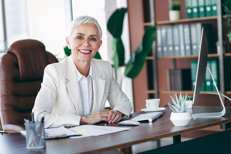 Elegant senior businesswoman smiling confidently at her modern office desk with a professional and stylish demeanorの写真素材